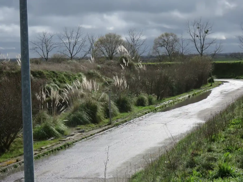 Route étroite longeant un fossé, talus sous un ciel nuageux.